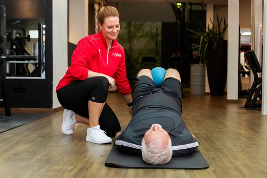 Fitnesscoach unterstützt männlichen Klienten bei einer Übung auf einer Matte in einem modernen Fitnessstudio.