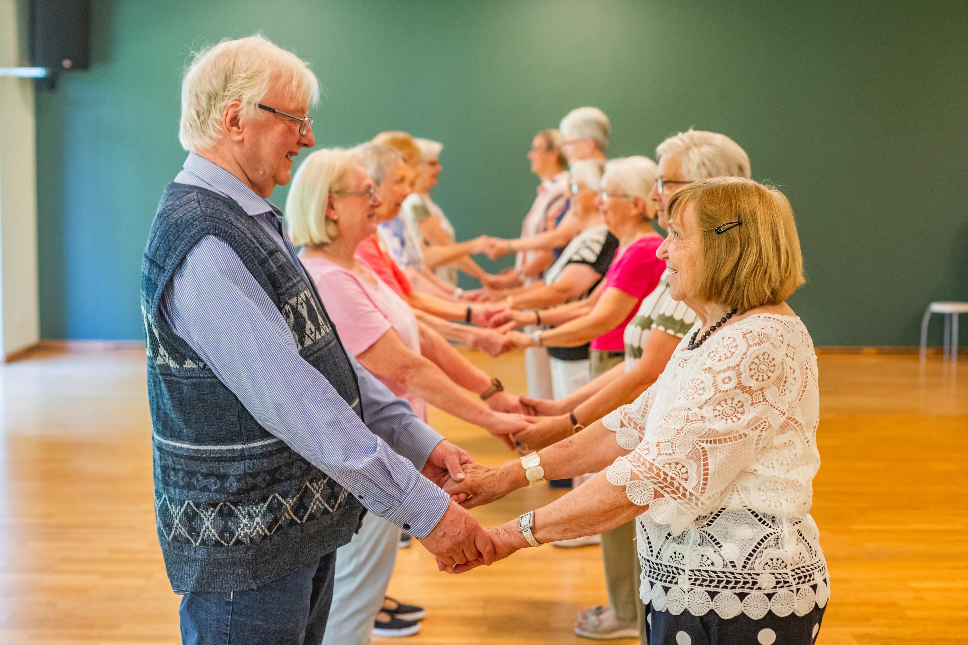 Ältere Menschen stehen in einer Reihe und halten sich an den Händen in einem lichtdurchfluteten Raum.